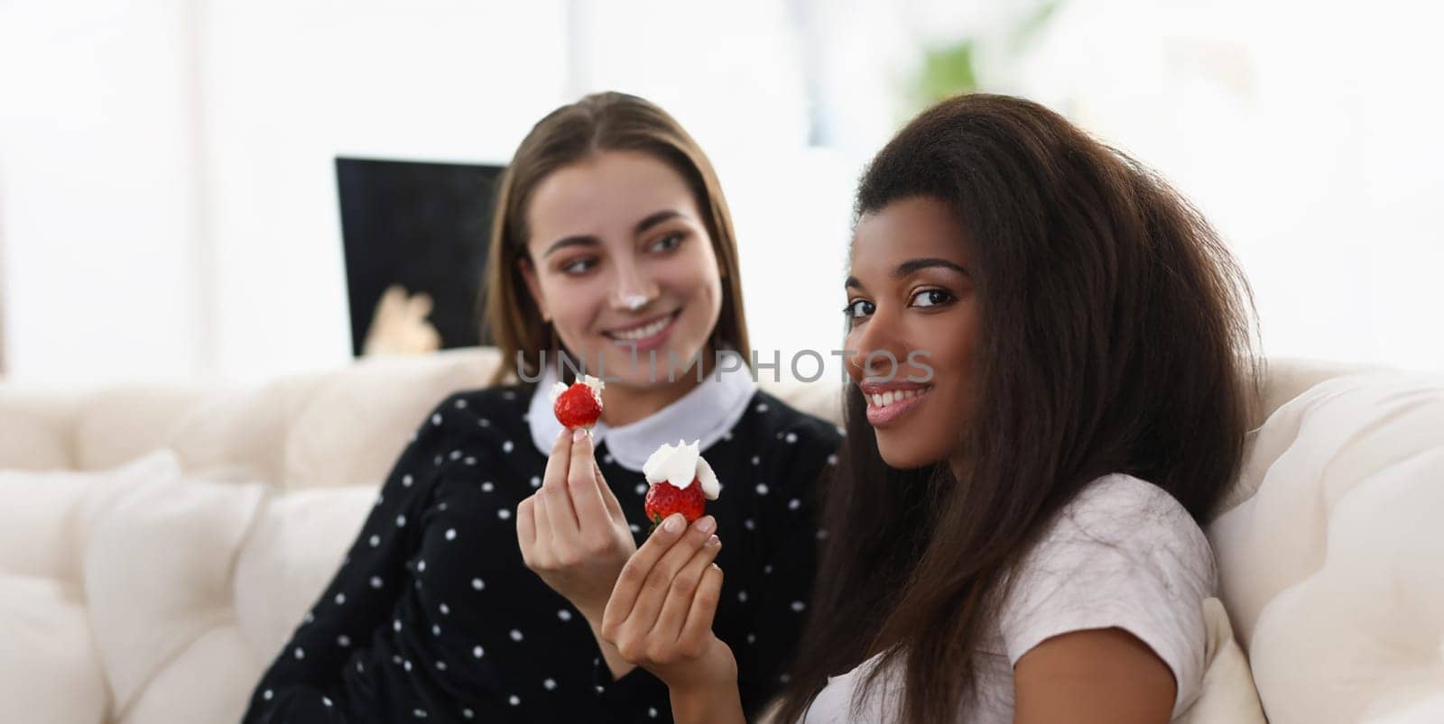 Portrait of smiling girls eating tasty fresh strawberry covered with whipped cream. Romantic date at home, best friends chilling. Lgbt, bff, lunch concept
