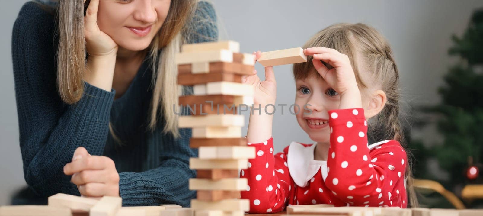 Portrait of pretty little kid pulling wooden block out of tower and smile. Mother join daughter in funny board game at home. Parenthood, childhood concept