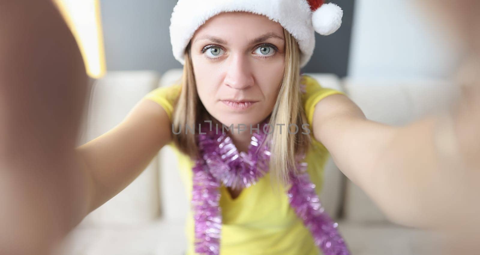 Portrait of beautiful serious blonde young woman posing on camera in santa claus hat. Celebrating new year alone at home. Holiday, christmas mood concept