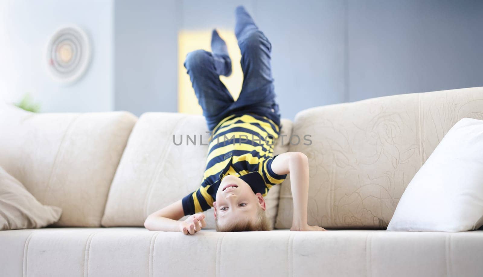 Portrait of young boy child standing on head on beige sofa going crazy at home on holiday. Alone at home, go nuts, lots of energy. Childhood, sport concept