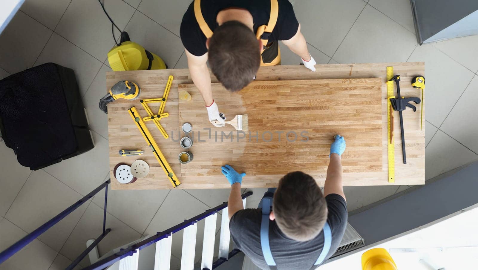Top view of coworkers covering wooden plank with shiny polish creative process in workshop. Male carpenters finish masterpiece in studio. Design concept