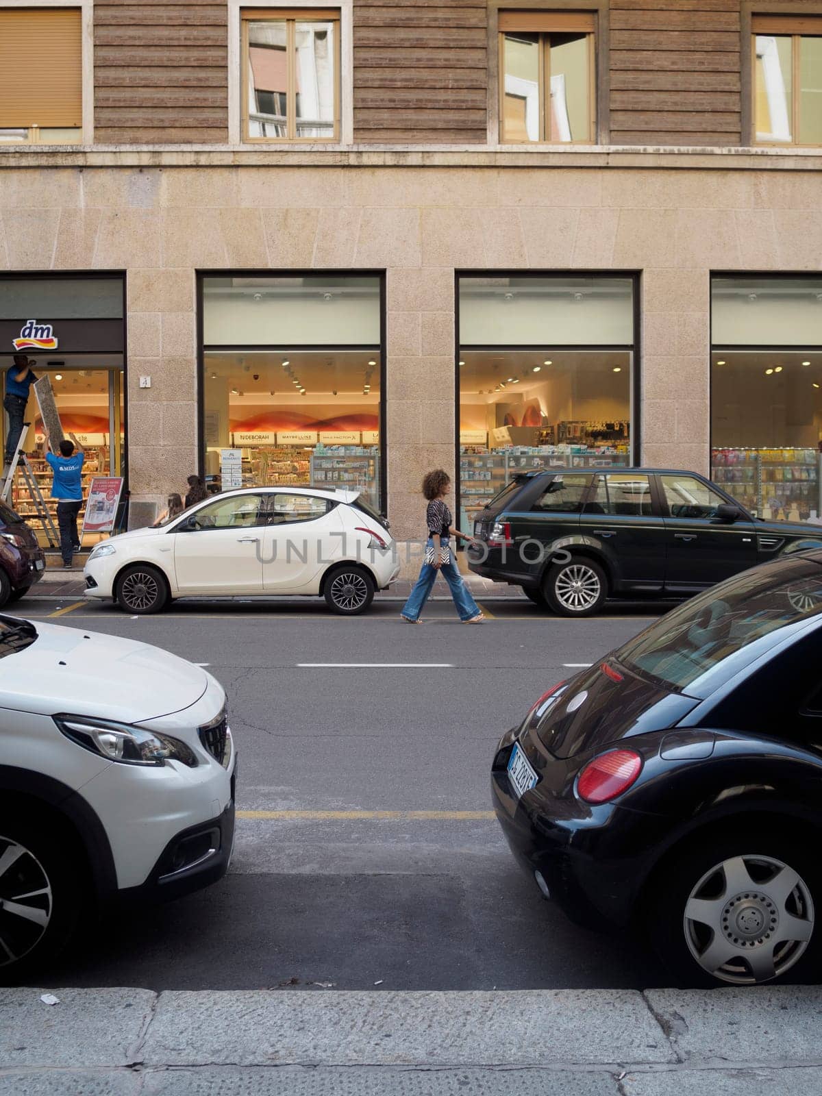 Cremona, Italy August 31st 2024 A woman is walking in a european city street with parked cars on the side and workers setting up a shop