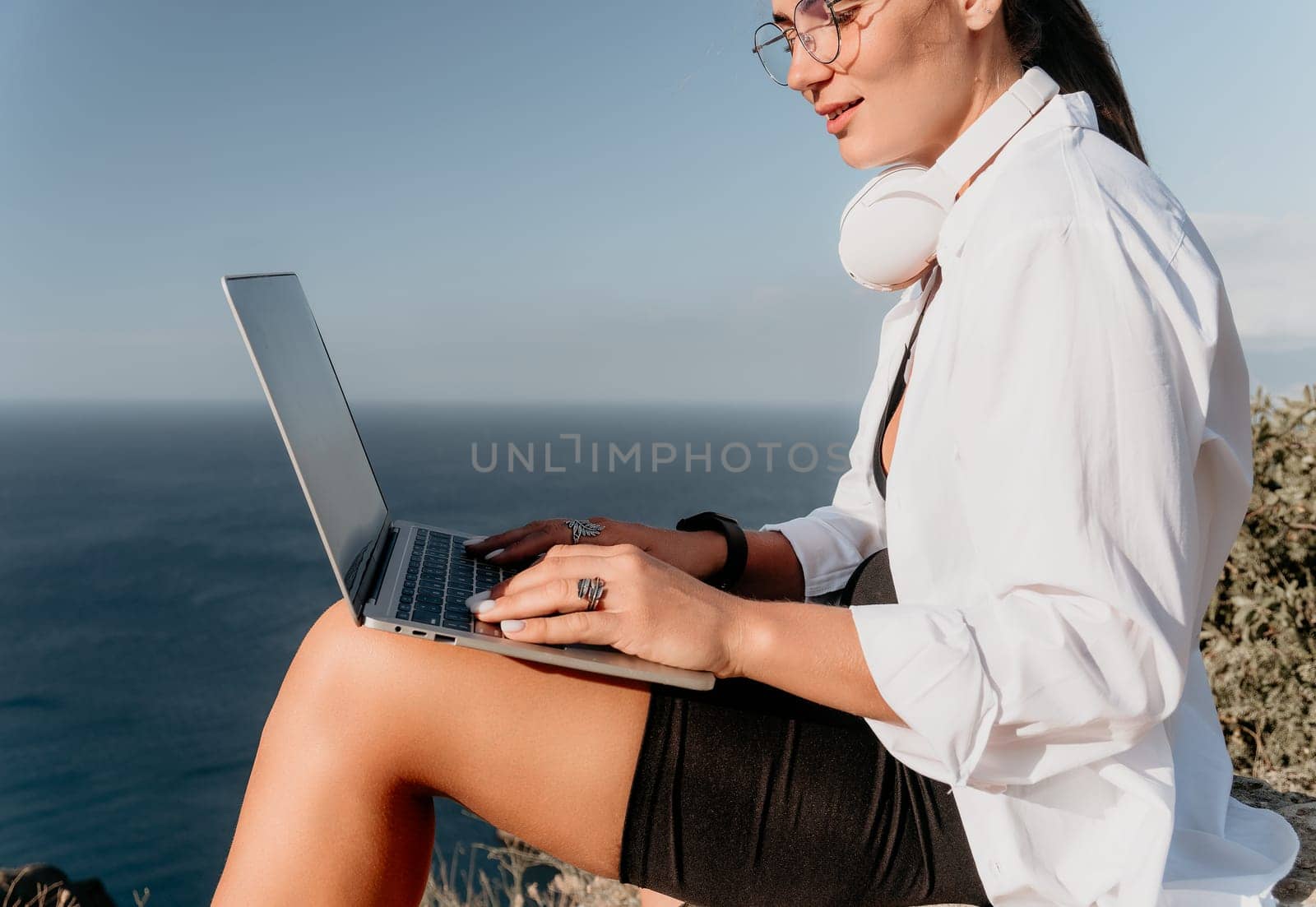 A woman wearing glasses and headphones sits on a cliff overlooking the ocean, typing on a laptop.