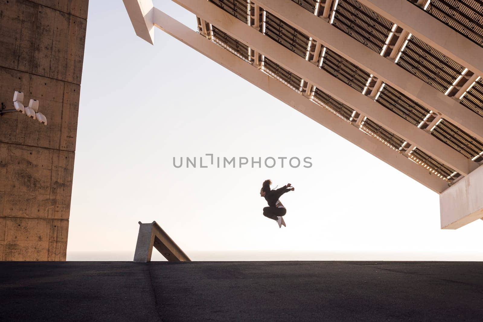 anonymous female athlete in sports clothes jumping with raised arms under modern construction with solar panels during workout, concept of urban sport and healthy lifestyle
