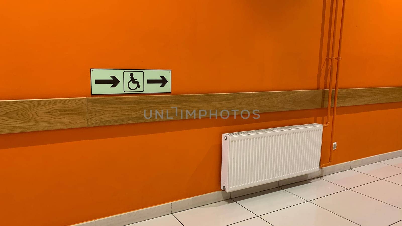 Corridor with orange wall in public place with direction sign for people on wheelchair with arrows, assistance in direction of equipped area with access for people with disabilities, way to wc