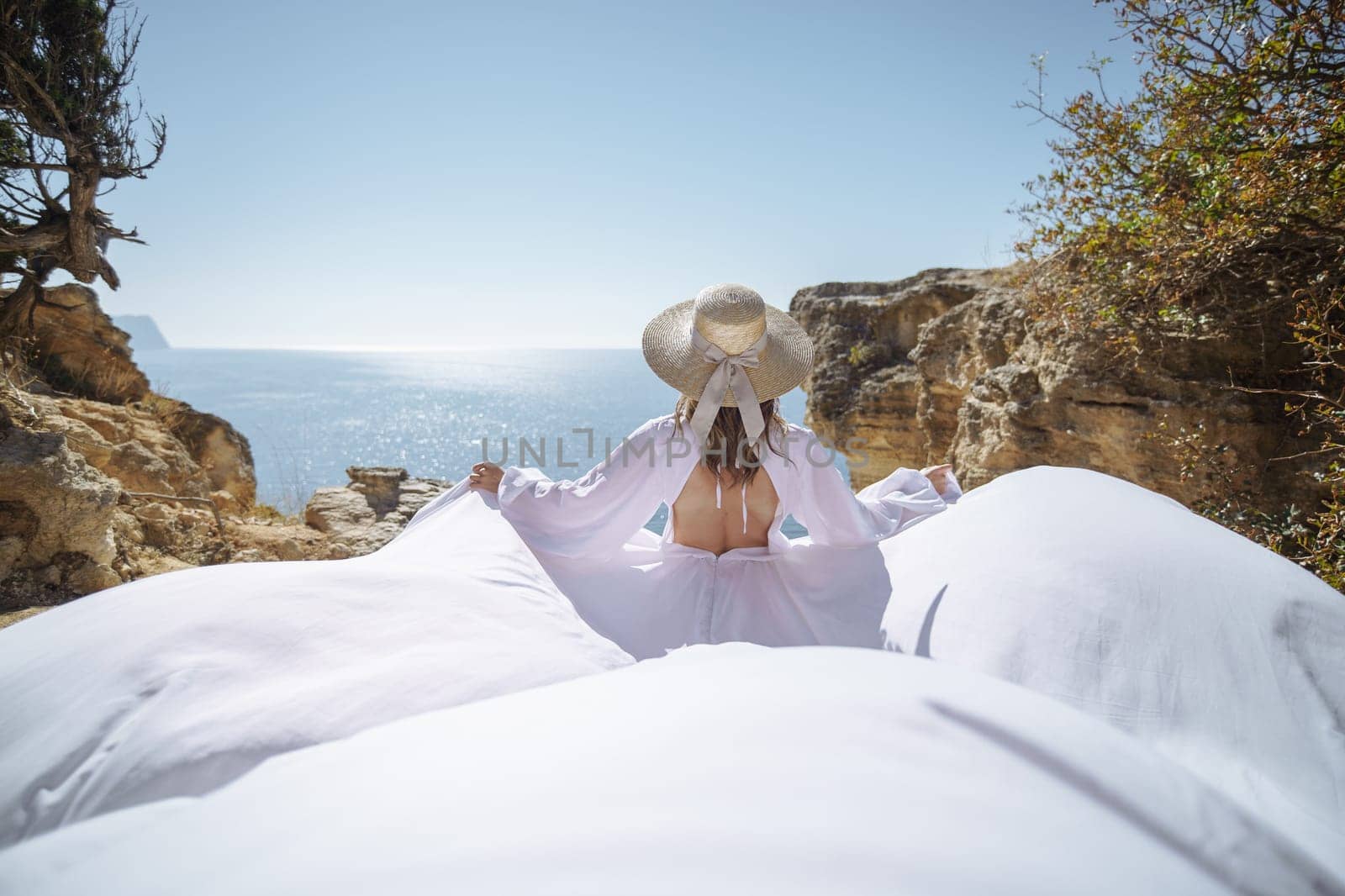 A woman in a white dress is standing on a rocky beach with her hat on. The scene is serene and peaceful, with the woman enjoying the view of the water.
