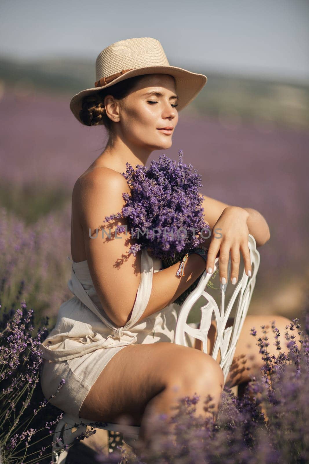 A woman is sitting in a field of lavender flowers and wearing a straw hat. She is smiling and holding a bouquet of flowers. Scene is peaceful and serene, as the woman is surrounded by the beauty of nature.