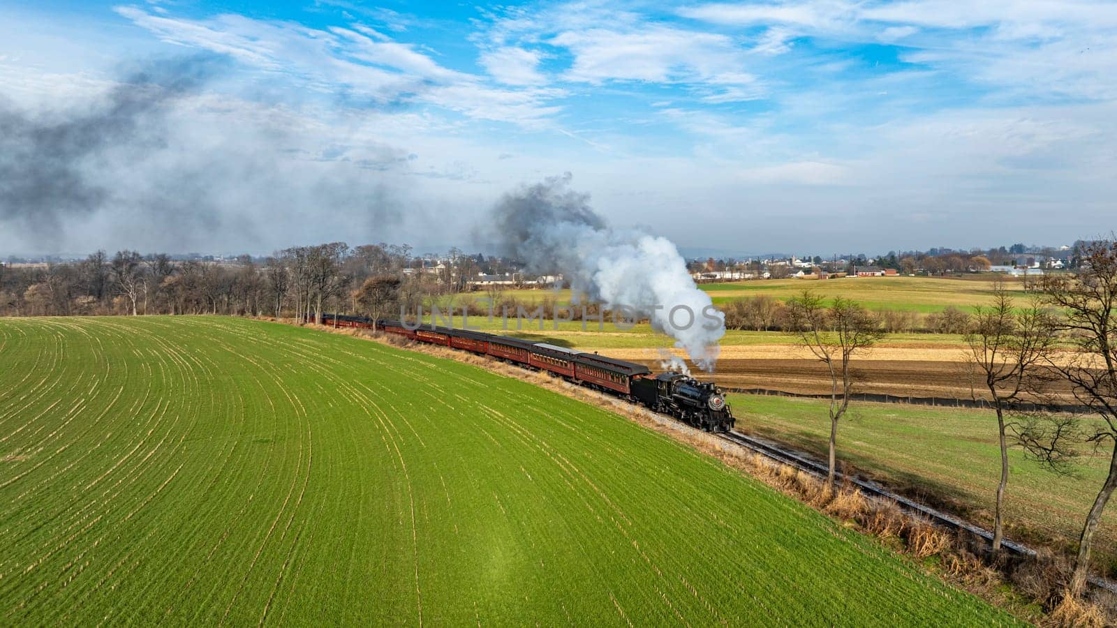 A charming steam train billows white smoke as it chugs along a picturesque landscape with vibrant green fields and a clear blue sky. . High quality photo