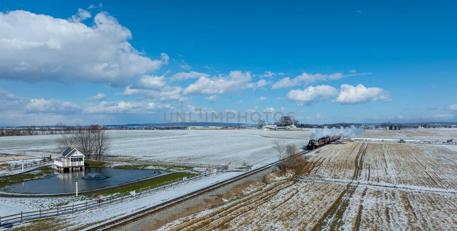 A steam locomotive chugs along the tracks, releasing vapor as it moves through snow-covered fields. In the foreground, a gazebo stands near a small pond, contributing to the serene winter landscape