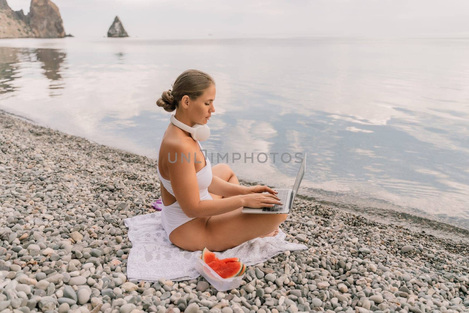 A woman is sitting on the beach with a laptop open in front of her. She is wearing a white bikini and has a pink headband on. Concept of relaxation and leisure