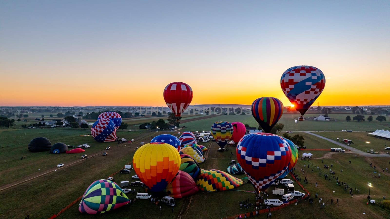 A dozen colorful hot air balloons are preparing for launch in a festival field during sunset. Spectators gather to witness the spectacle as the evening sky radiates warm colors.