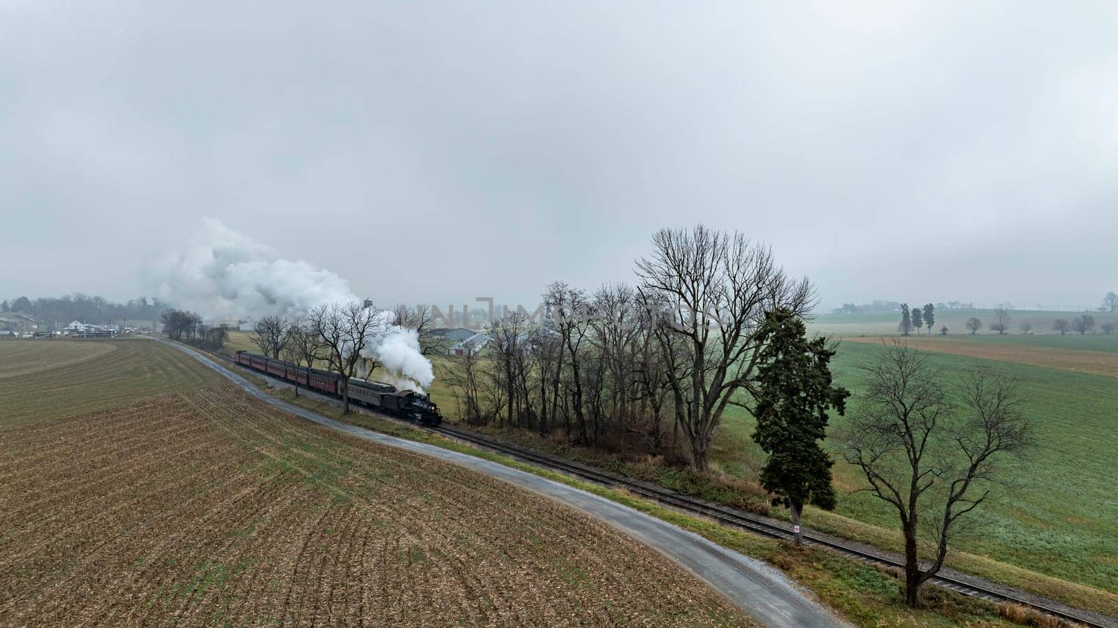 A vintage steam train glides along railway tracks bordered by fields and trees under a cloudy, fog-laden sky. The landscape showcases a serene rural environment in early morning light.