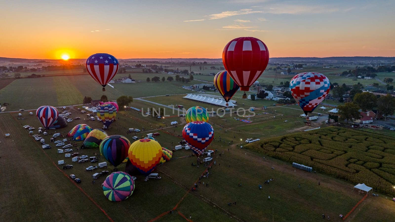 A dozen colorful hot air balloons are preparing for launch in a festival field during sunset. Spectators gather to witness the spectacle as the evening sky radiates warm colors.