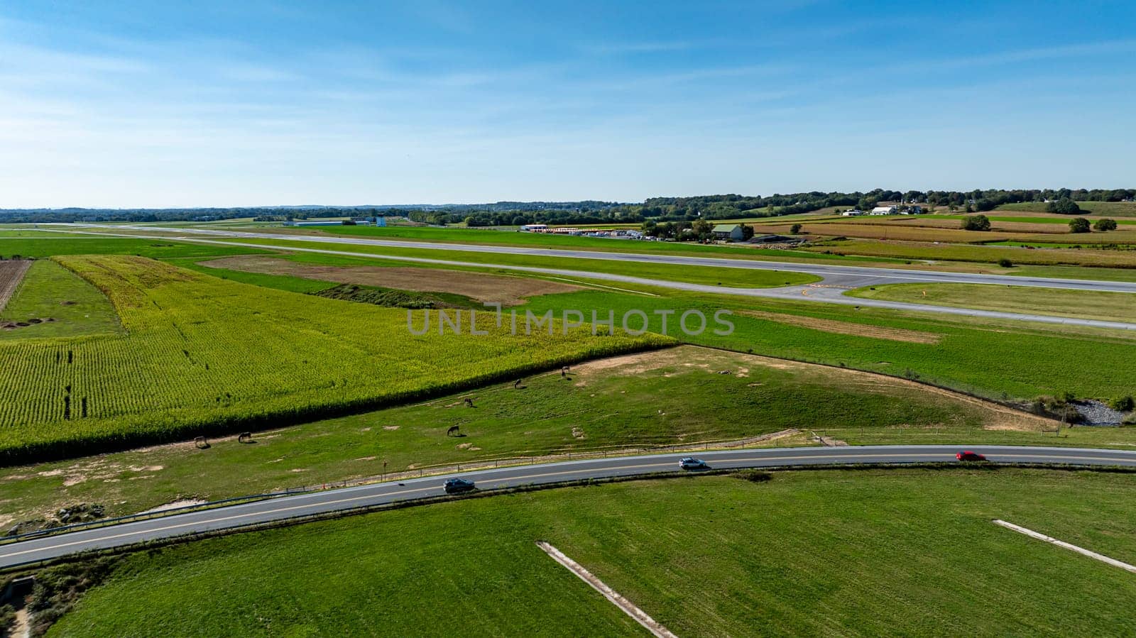An expansive view showcases vibrant green fields of farmland alongside a small airstrip. The landscape is serene, with a clear blue sky enhancing the rural charm of the area.