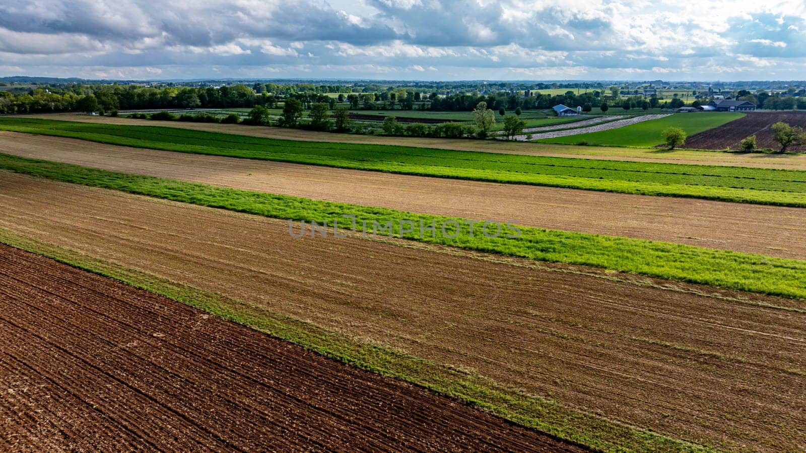 Expansive farmland stretches across the landscape, featuring alternating crop rows in varying shades of green and brown. Cloudy skies hover above the rich agricultural fields.
