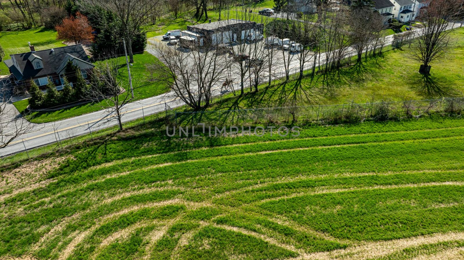 The scene captures an aerial perspective of a vibrant green field marked by gentle curves, adjacent to a winding road. Nearby, a blend of homes and vehicles adds to the rural charm of the area.