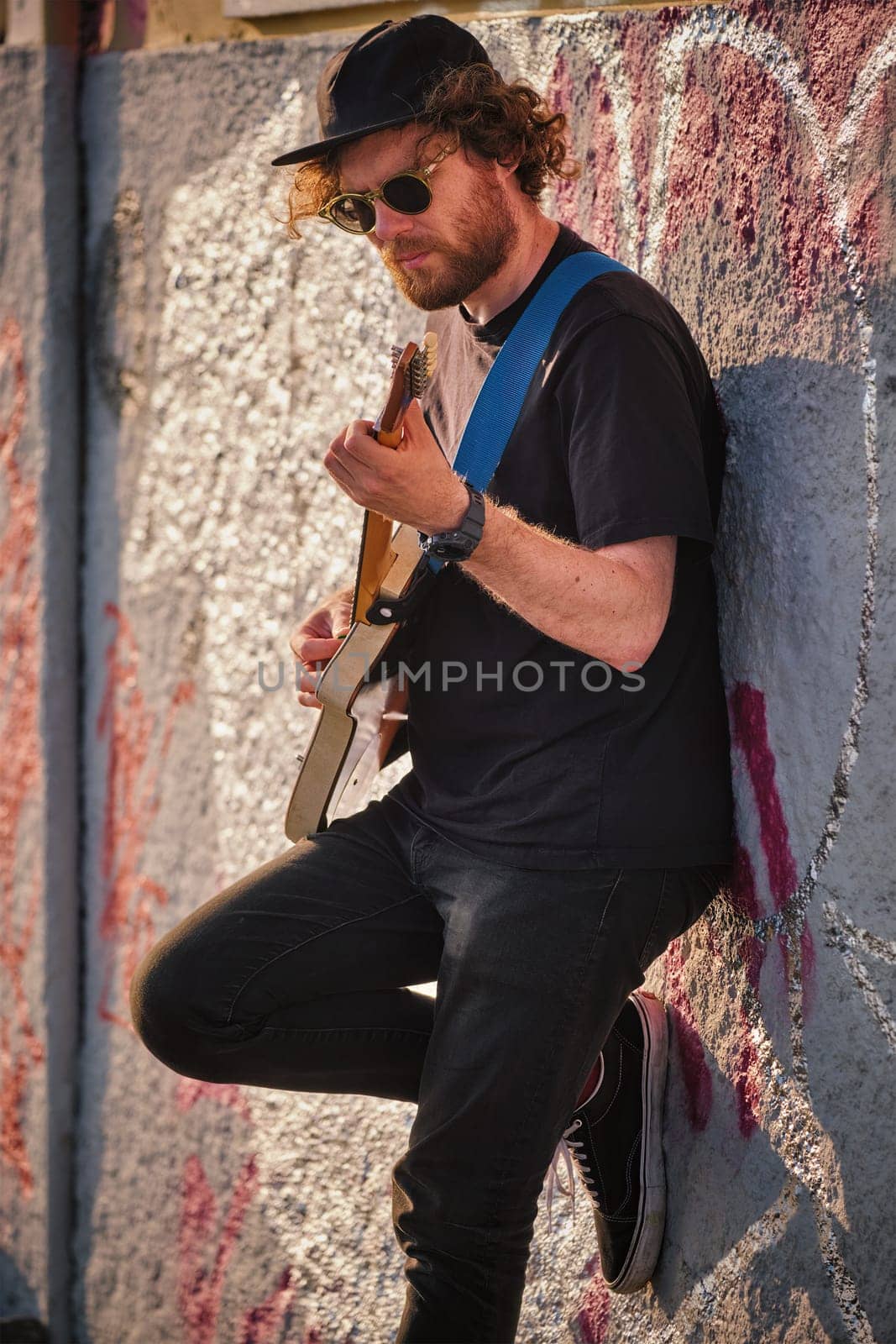 Hipster street musician in black playing electric guitar in the street on sunset leaning on a wall. Lisbon, Portugal