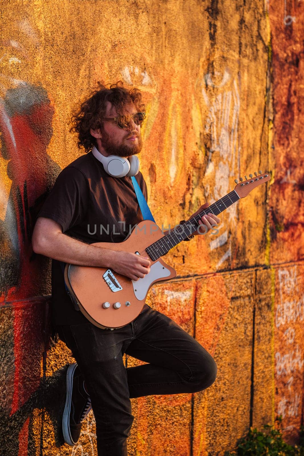 Hipster street musician in black playing electric guitar in the street on sunset leaning on a painted wall