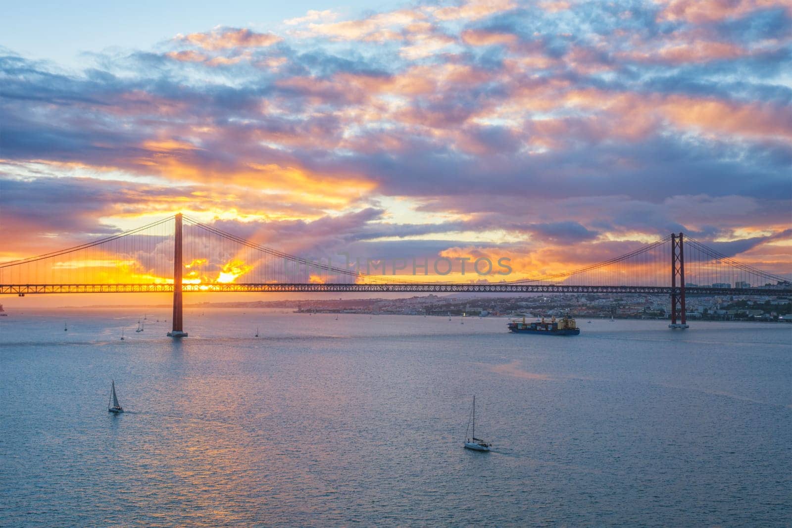 View of 25 de Abril Bridge famous tourist landmark of Lisbon over Tagus river with tourist yacht boats and cargo container ship on sunset. Lisbon, Portugal