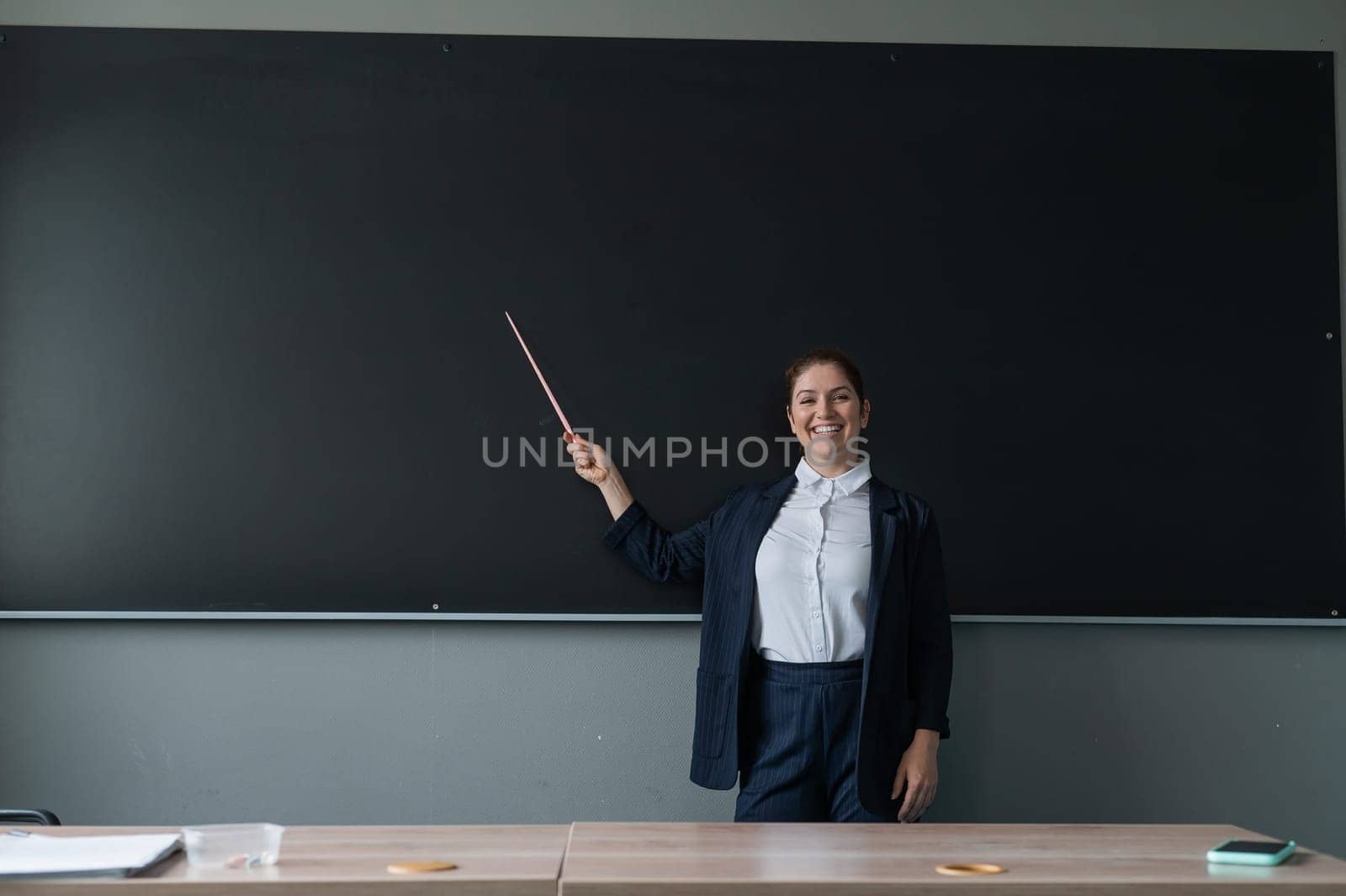 Red-haired caucasian woman in a trouser suit. Smiling female teacher with a pointer at the blackboard