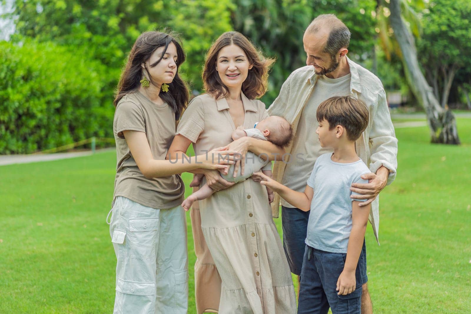 Happy couple in their 40s, surrounded by adult children, cradle a newborn in a sunlit park. Love, family, and generations in harmony.