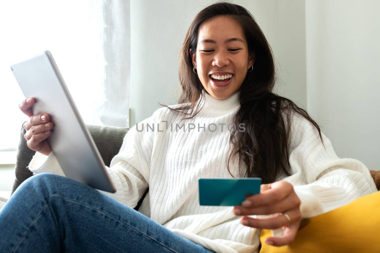 Happy young Asian woman using digital tablet and credit card to shop online sitting on the sofa at home living room. Copy space. E-commerce concept.