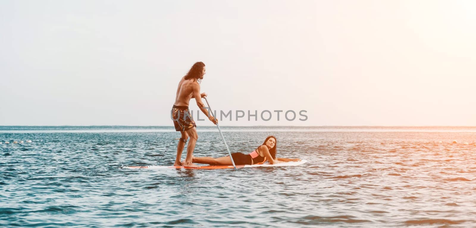 Sea woman and man on sup. Silhouette of happy young woman and man, surfing on SUP board, confident paddling through water surface. Idyllic sunset. Active lifestyle at sea or river. by panophotograph