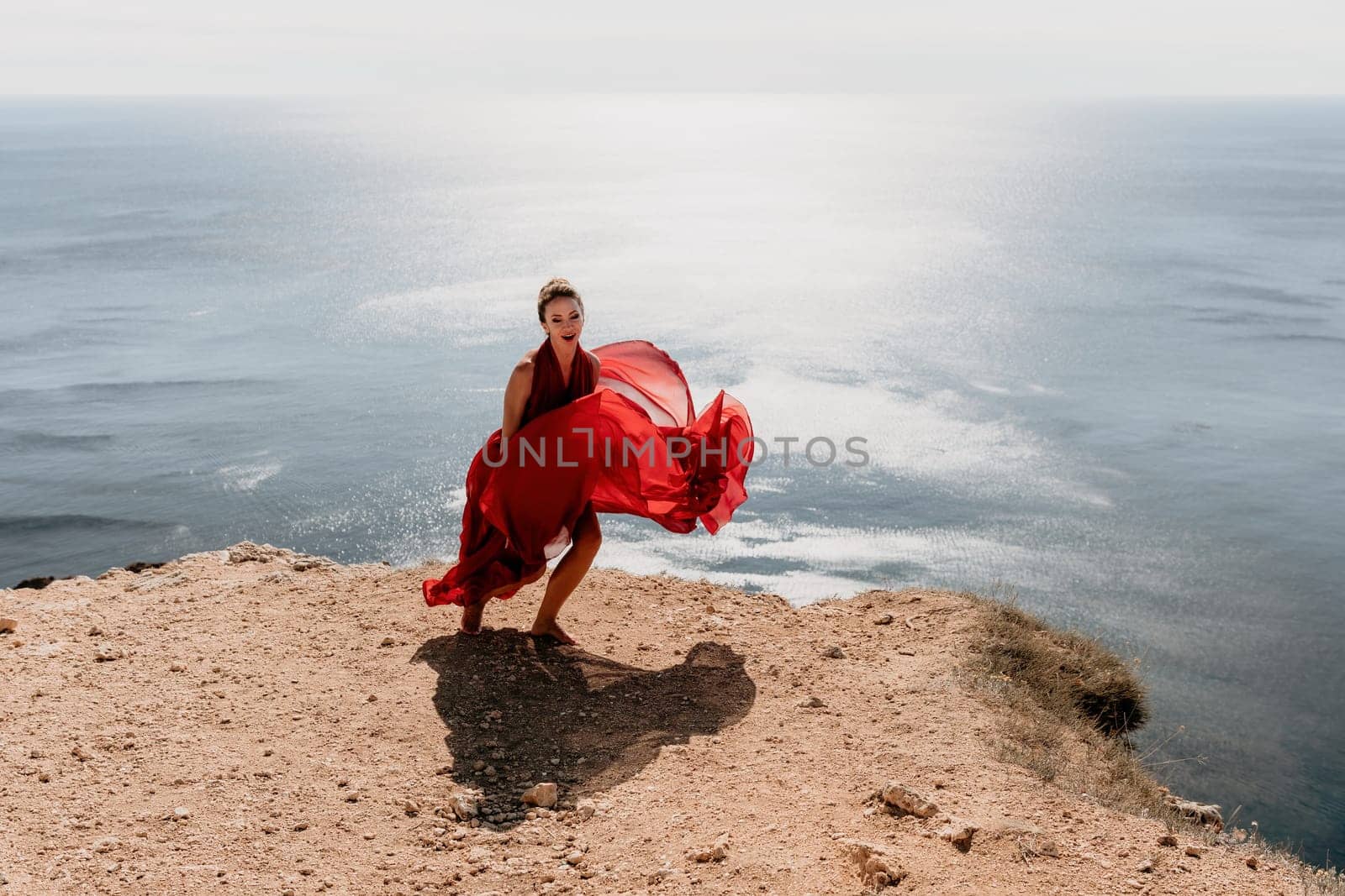 Side view a Young beautiful sensual woman in a red long dress posing on a rock high above the sea during sunrise. Girl on the nature on blue sky background. Fashion photo.