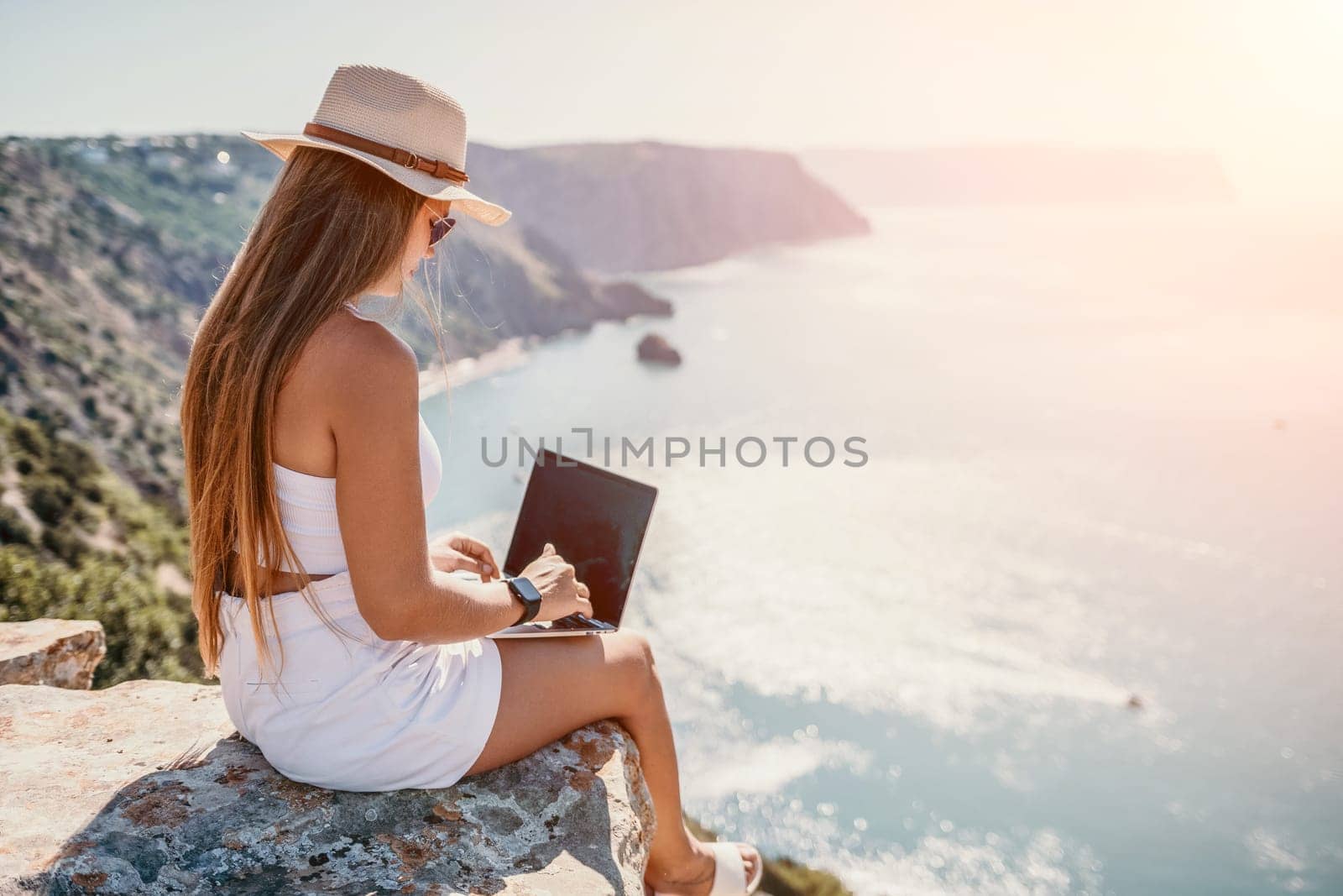 Successful business woman in yellow hat working on laptop by the sea. Pretty lady typing on computer at summer day outdoors. Freelance, travel and holidays concept.