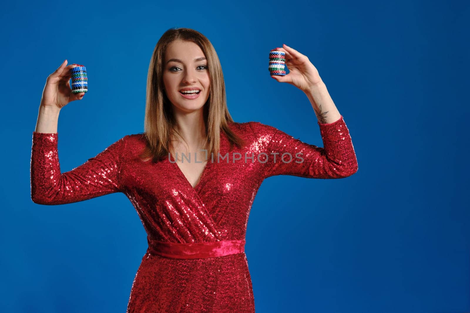 Beautiful blonde model with make-up, tattooed hand, in red sequin dress is smiling, showing some colorful chips, posing against blue background. Gambling entertainment, poker, casino. Close-up.