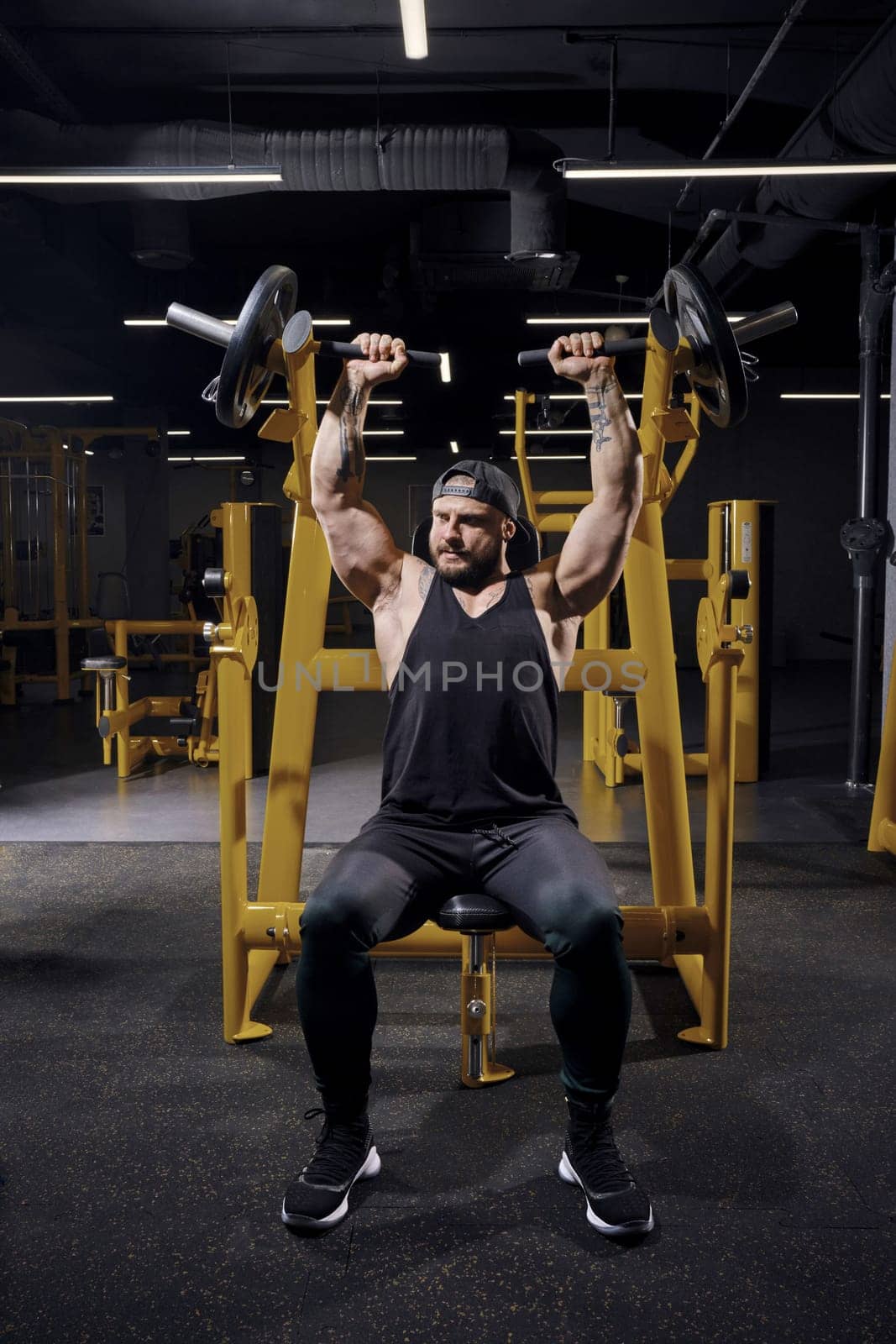 Tattooed, bearded male in black sweatpants, vest and cap. He performing a chest press while sitting on an exercise machine at dark gym. Full length by nazarovsergey