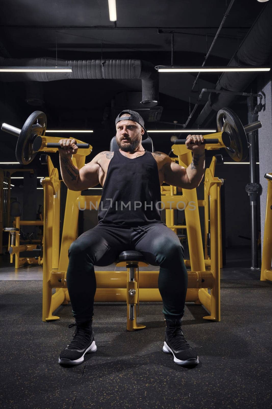 Tattooed, bearded, strong guy in black sweatpants, vest and cap. He performing chest press, sitting on an exercise machine at dark gym. Full length by nazarovsergey