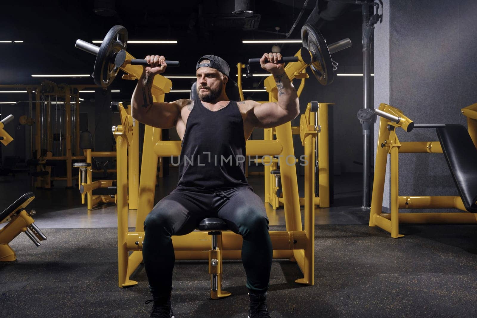 Tattooed, bearded sportsman in black sweatpants, vest and cap. He performing a chest press while sitting on an exercise machine at dark gym. Close up by nazarovsergey