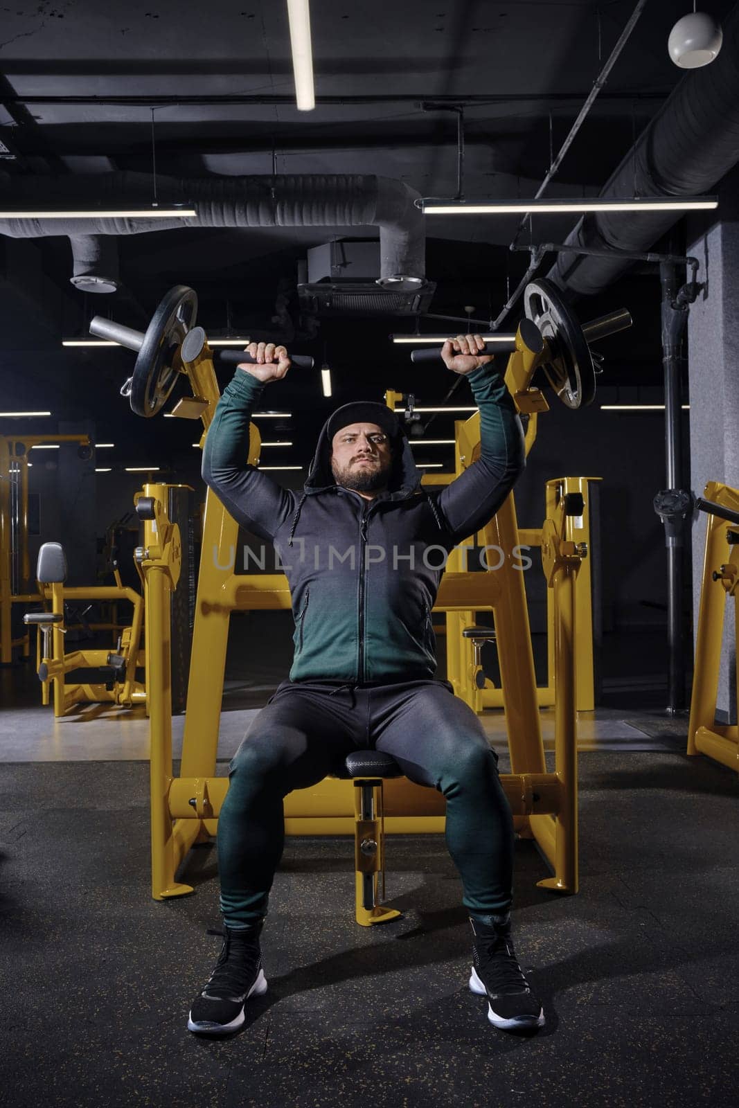 Bearded man in black tracksuit with a hood, cap and sneakers. Performing chest press, sitting on an exercise machine, posing in dark gym. Full length by nazarovsergey