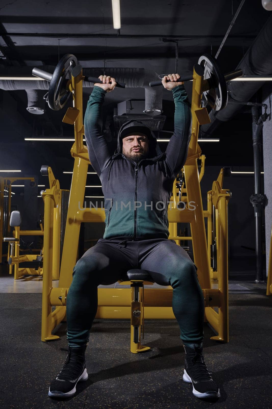 Muscular bearded male in black tracksuit with a hood, cap and sneakers. He performing chest press, sitting on an exercise machine, posing in dark gym with yellow equipment. Sport, fitness. Full length