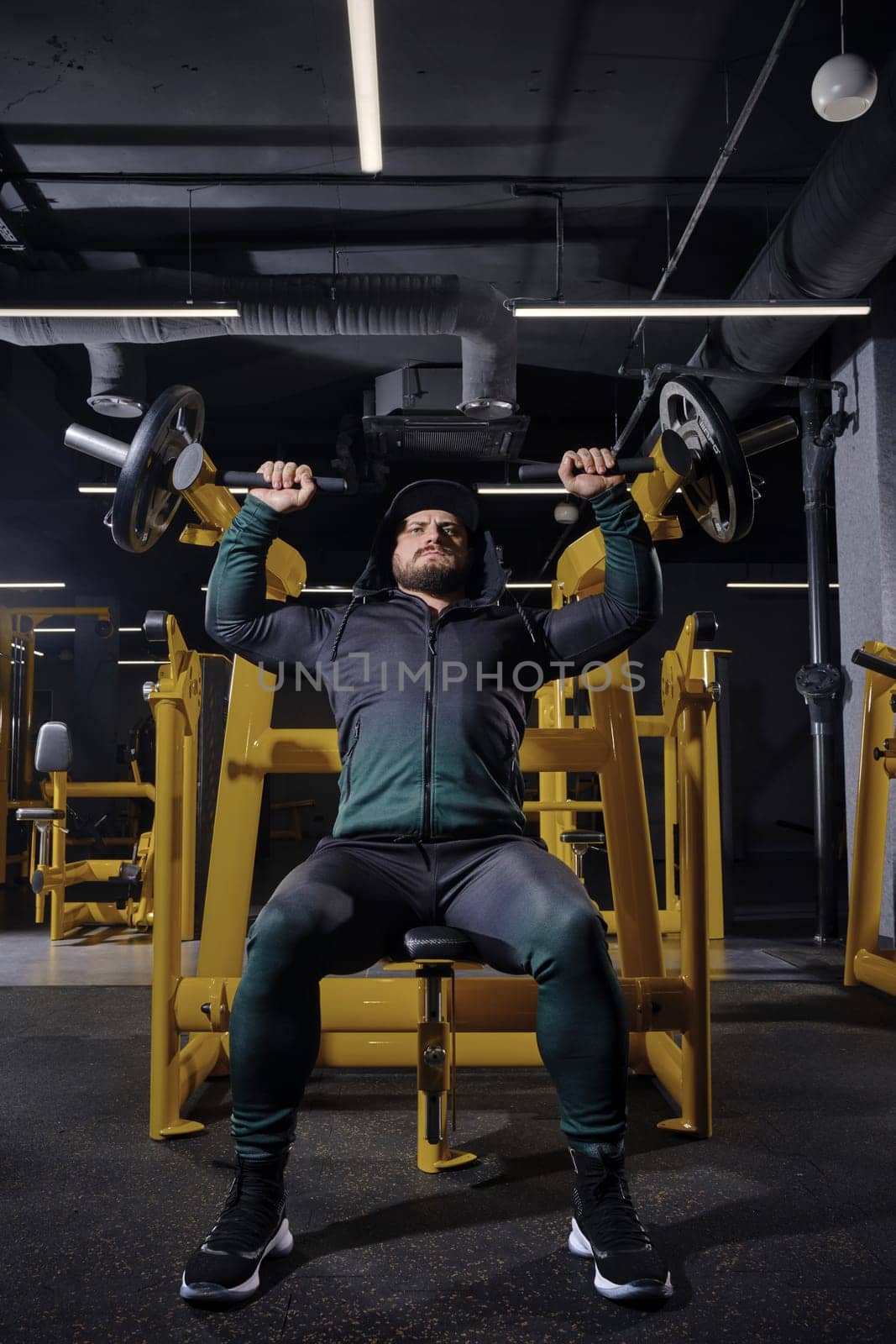 Attractive, bearded sportsman in black tracksuit with a hood, cap and sneakers. He performing a chest press while sitting on an exercise machine, posing in dark gym with yellow equipment. Full length