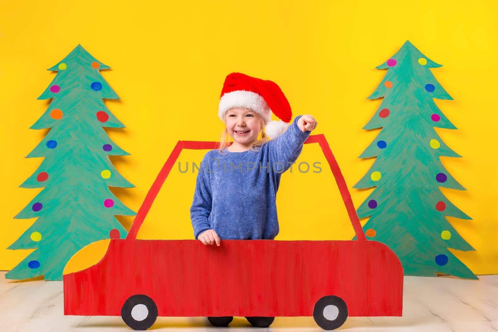 Child with Christmas hat driving a car made of cardboard. Little girl having fun at home on a yellow background. Christmas concept. New Year's holidays.