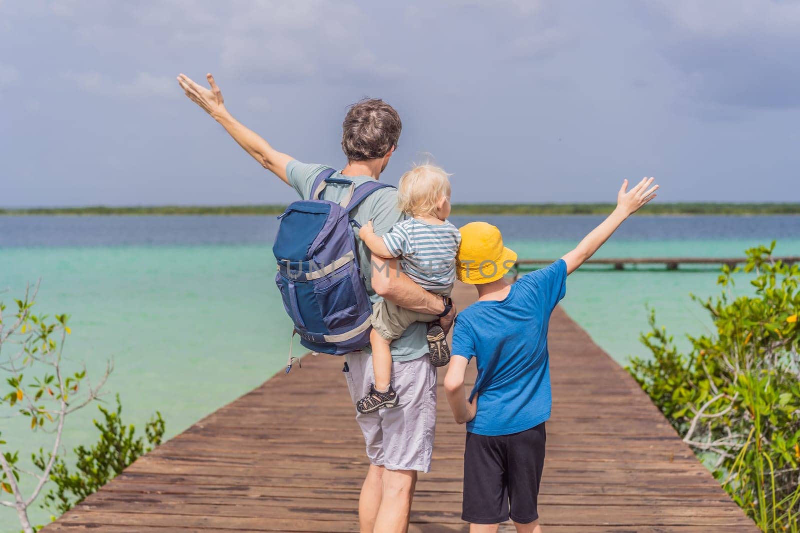 Father with his toddler and teenage sons exploring wooden pier over the turquoise waters of Bacalar Lake, Mexico. Peaceful tropical travel destination concept.