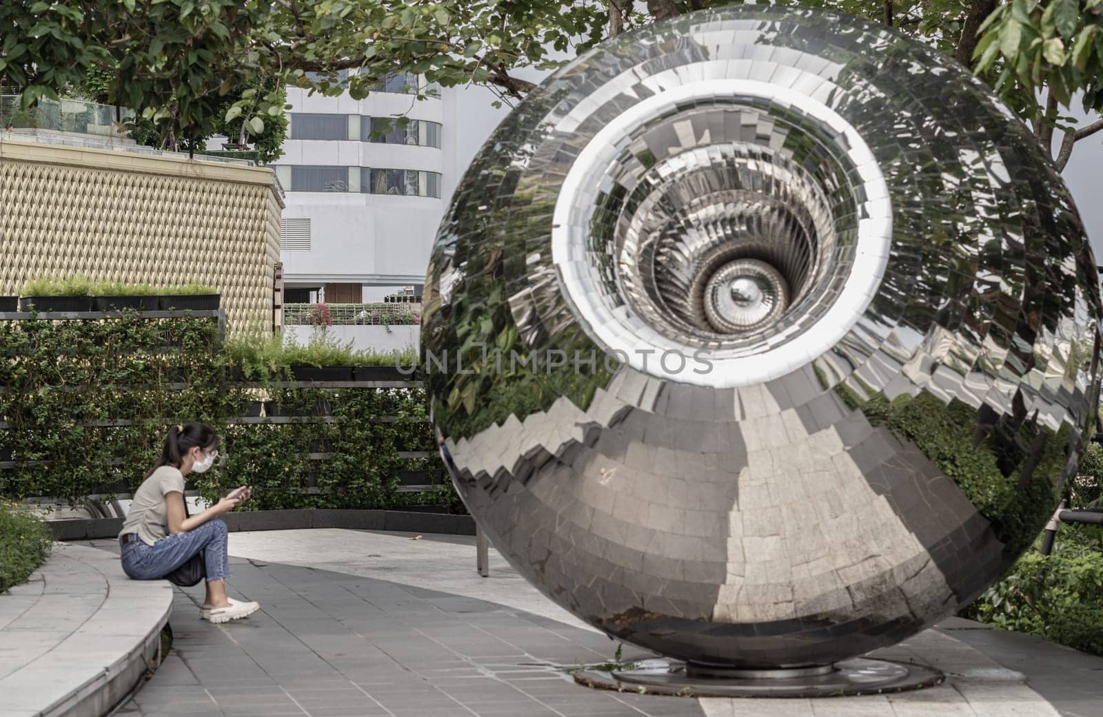 Bangkok,​ Thailand - 28 Sep, 2024 - An unidentified young woman wearing egg-colored t-shirt with jeans and protective face mask and using a smartphone while sitting alone sit rest in city park with Stainless steel pupil shape sphere sculptures, Space for text, Selective focus.