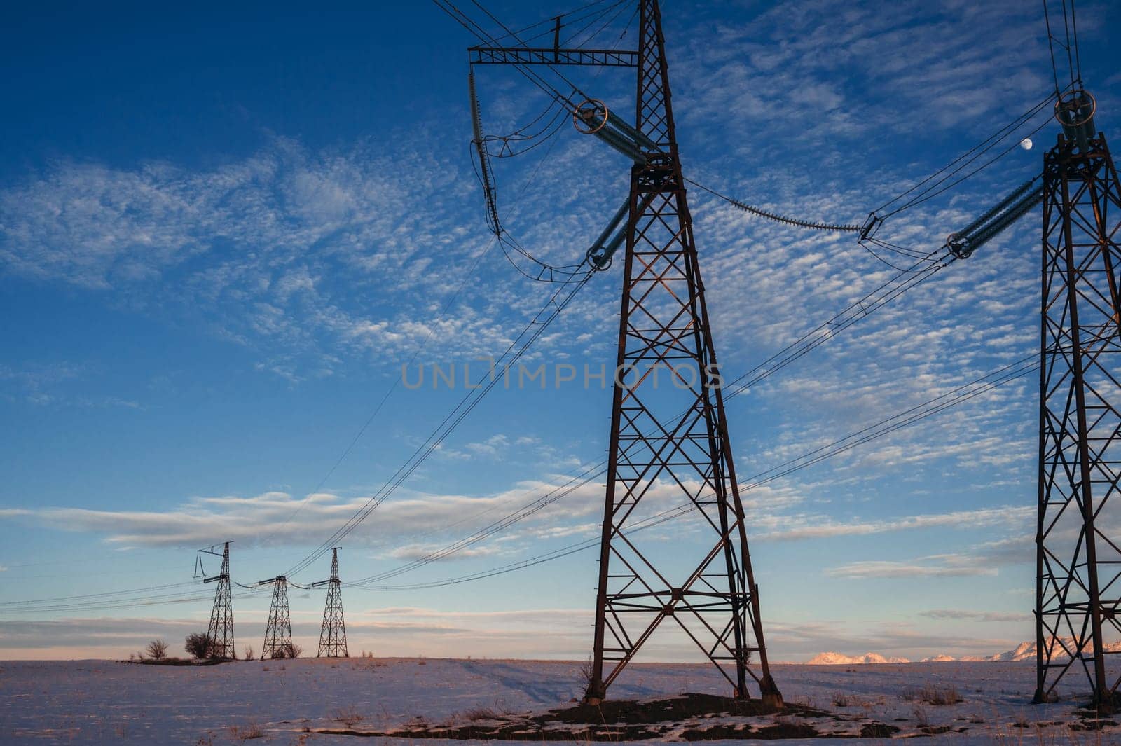 High-voltage power transmission line with towers in the field in winter at sunset in the evening