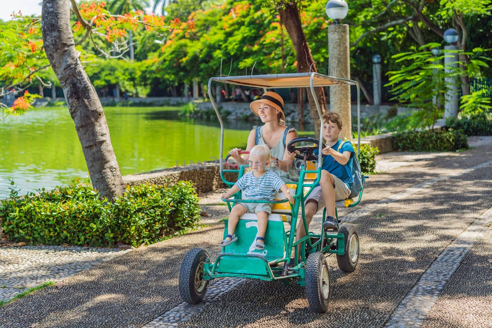Mother tourist with her toddler and teenage sons riding an authentic Mexican pedal car in a park in Mexico. Family adventure and travel with kids concept.