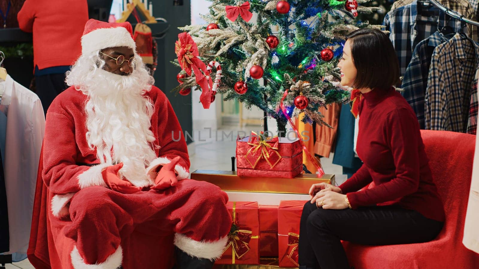 Asian woman engaging in conversation with employee impersonating Santa Claus during festive Christmas shopping experience. Customer receiving xmas present from worker in fashion shop