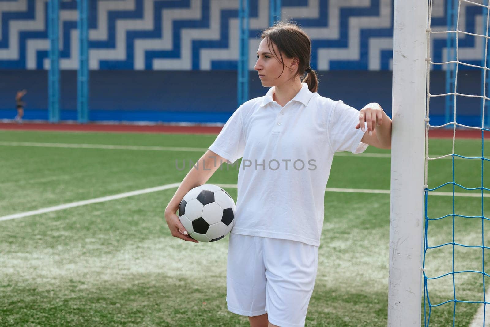 Female soccer player in white uniform holding a soccer ball and leaning against a goalpost on a soccer field.
