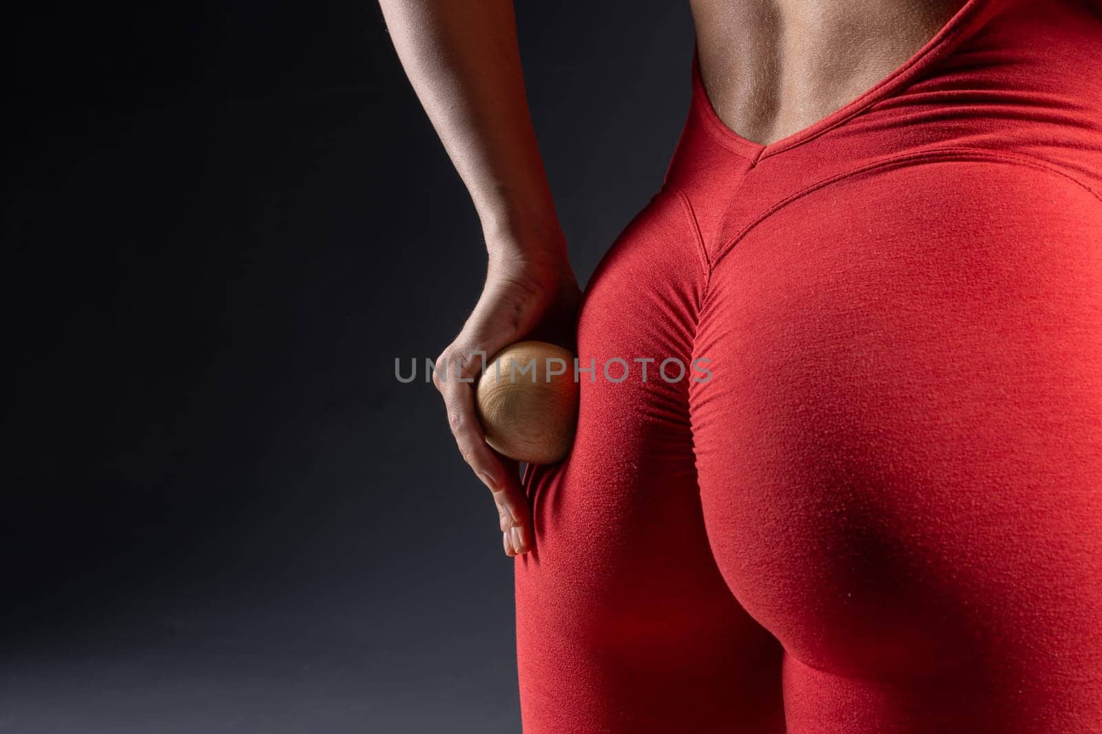 Close up of woman using wooden yoga massage ball on buttock in red tank top on dark background, self care and relaxation techniques. Self massage tool, cellulite removal concept