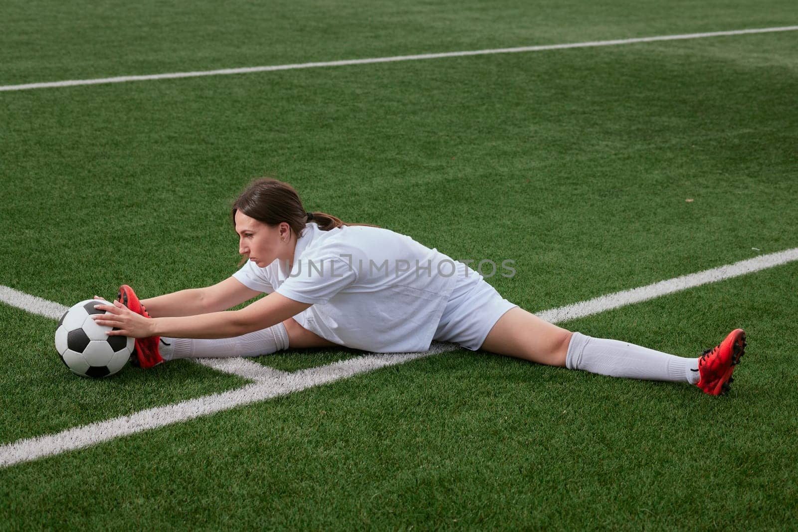A young soccer player stretches on a green field. She is in a split position, reaching for a soccer ball with both hands. Preparing for a game.
