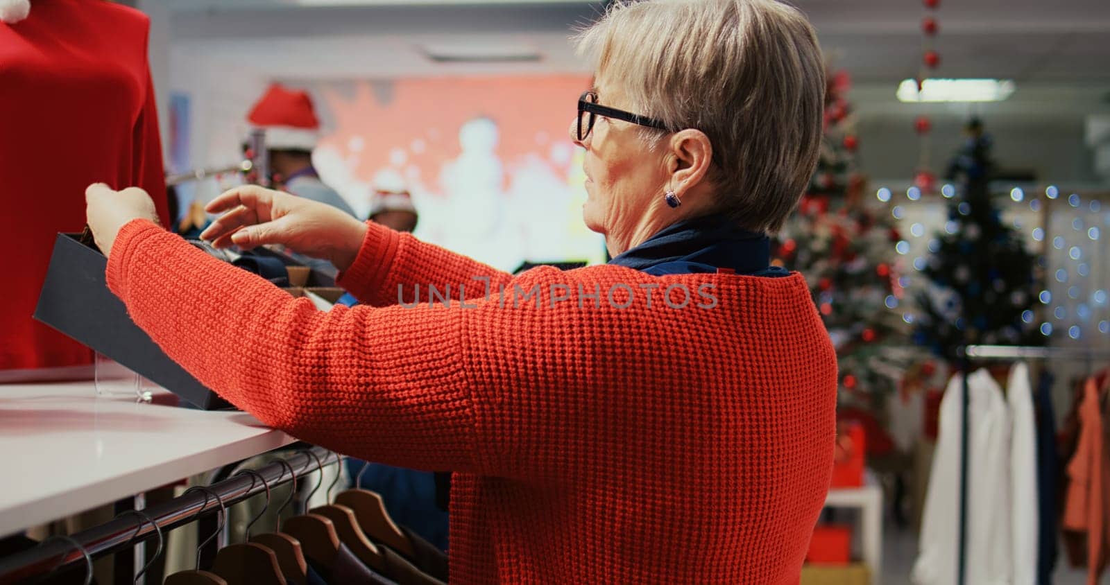 Elderly woman shopping for stylish neckties to gift husband for Christmas during winter holiday season. Senior customer in xmas decorated clothing store looking for presents