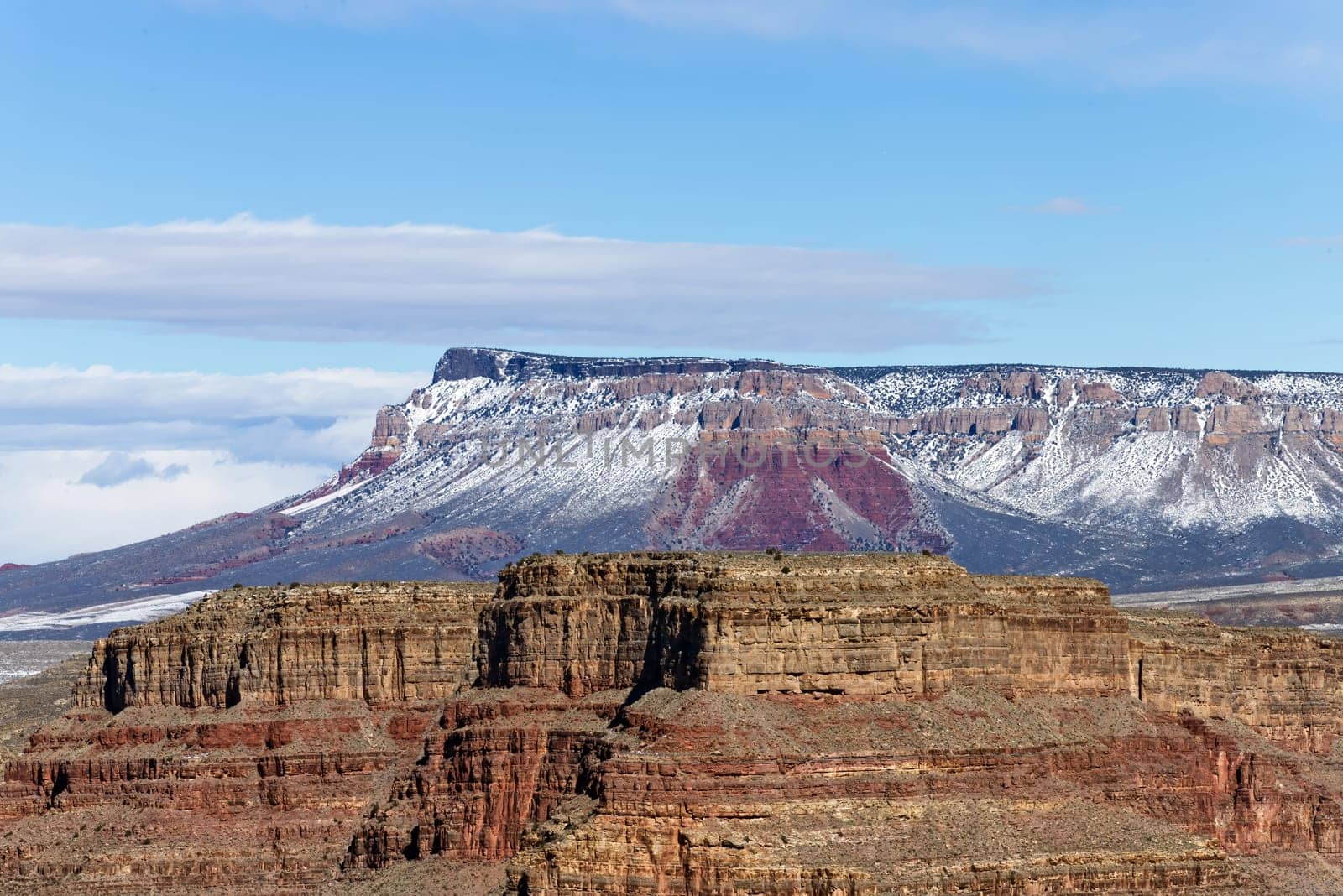 Experience ethereal beauty with this high-resolution photograph showcasing Grand Canyon West set against a sky gently kissed by clouds. The image captures the canyon's rugged red-rock formations reaching toward the heavenly horizon, as wisps of clouds float by, adding a dreamy softness to the tableau. The radiant blue sky provides a striking contrast, elevating the scene to surreal heights.