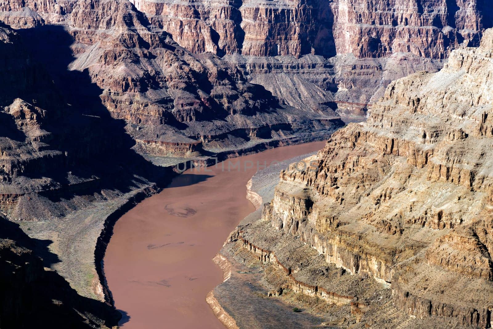 Witness the mighty Colorado River as it winds through the base of Grand Canyon West, captured at an elevation of 1,160 ft (350 m) in this high-resolution photograph. The image provides a mesmerizing perspective on the river role in carving this iconic landscape over millennia. The water emerald hue contrasts beautifully with the towering red-rock formations, creating a scene of breathtaking grandeur. Perfect for adding a sense of depth and history to your home, office, or digital project