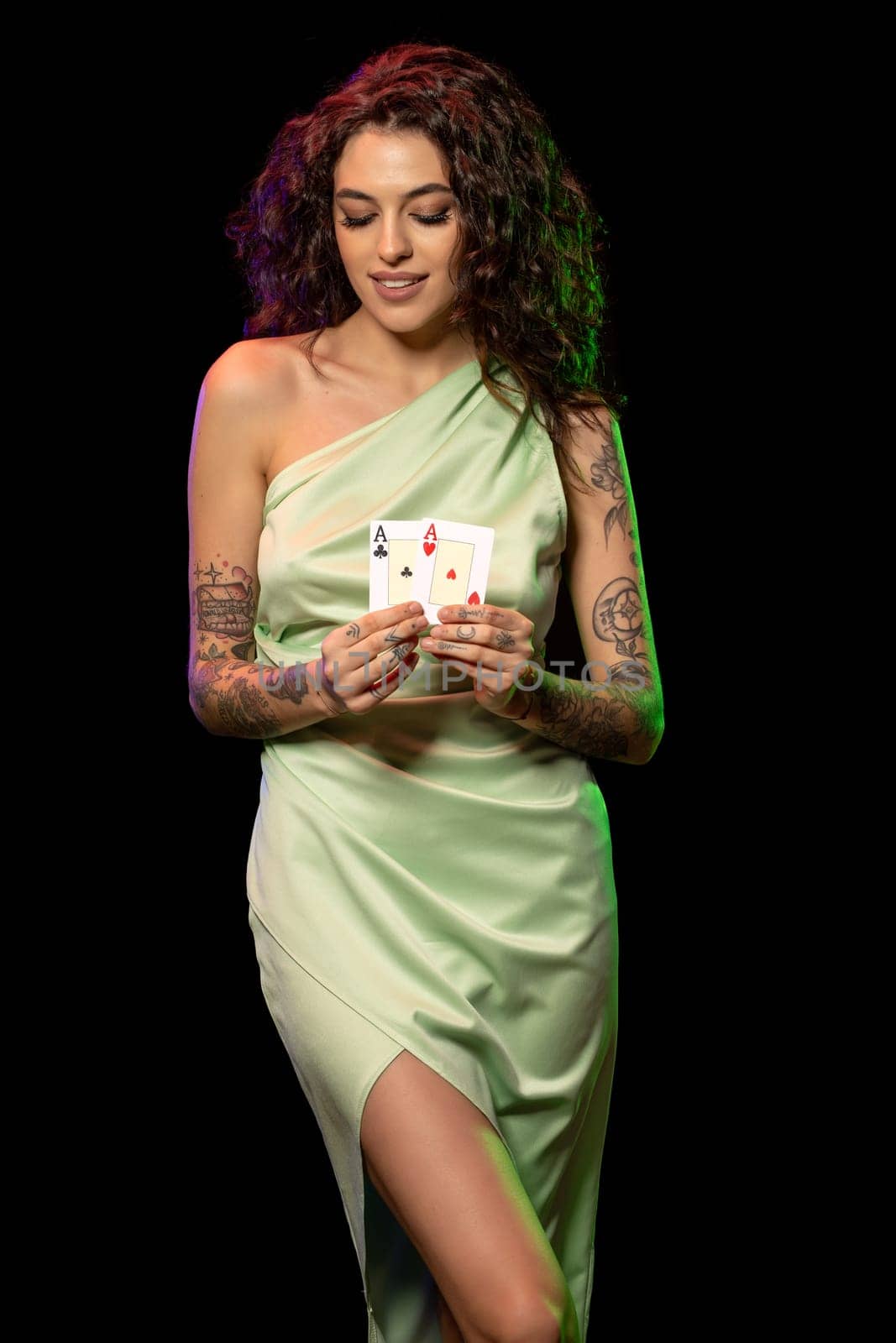 Lucky young brown-haired girl in elegant light green dress satisfied with successful poker game standing against black background, showing set of winning cards from pair of aces. Fortune smiling