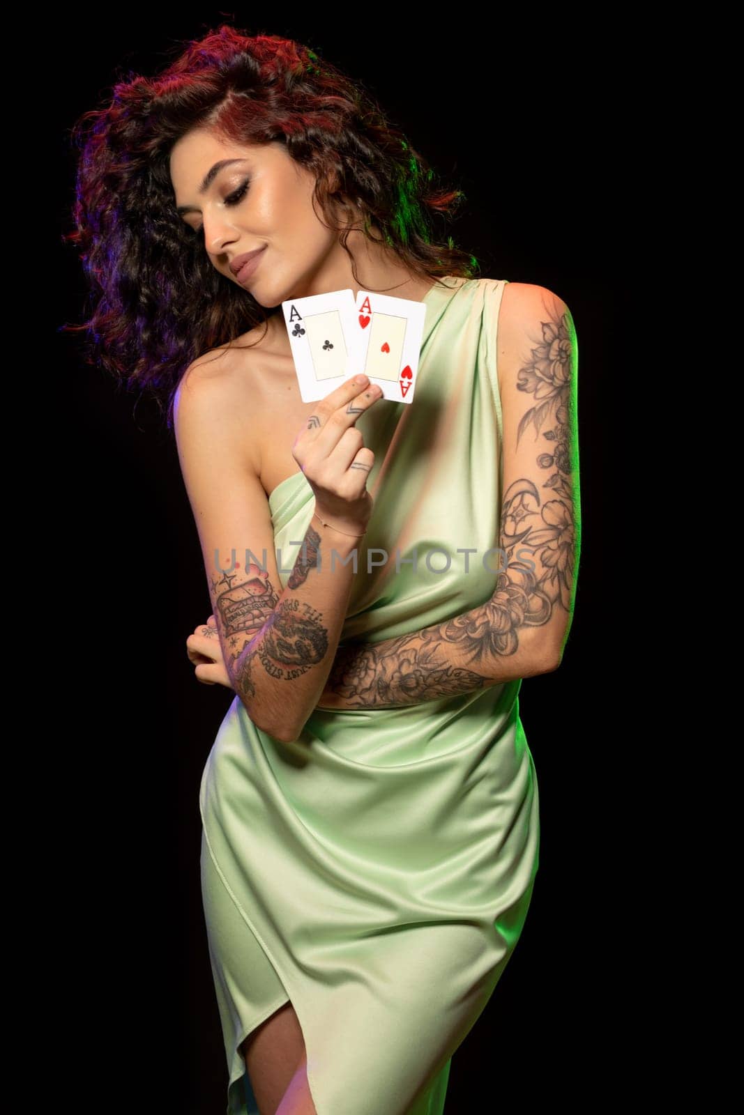 Portrait of smiling young woman in light green dress with tattoo on arms showing pair of aces, standing on dark studio background. Poker game concept